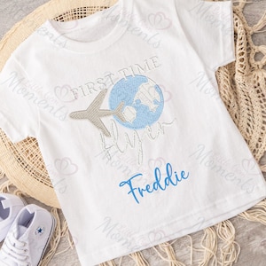 May include: White t-shirt with blue and gray embroidery of a globe, airplane, and the text "First Time Flyer". The name "Freddie" is embroidered in blue below the design.
