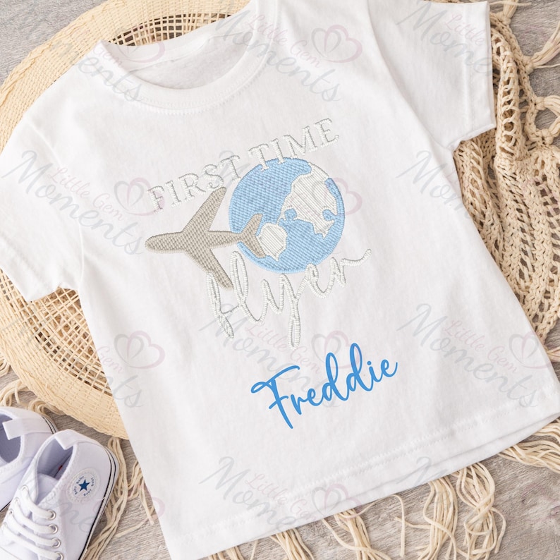 May include: White t-shirt with blue and gray embroidery of a globe, airplane, and the text "First Time Flyer". The name "Freddie" is embroidered in blue below the design.
