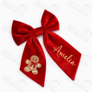 May include: A vibrant red velvet hair bow, featuring a gingerbread man and the name "Amelia" in gold lettering. This festive accessory is ideal for Christmas or seasonal celebrations.