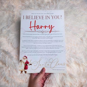 Puede incluir: Una carta personalizada de Papá Noel, impresa en papel blanco. El texto dice "I BELIEVE IN YOU!" con el nombre "Harry" en escritura roja. Una pequeña ilustración de Papá Noel está en la parte inferior, con las palabras "Santa Claus" en oro.