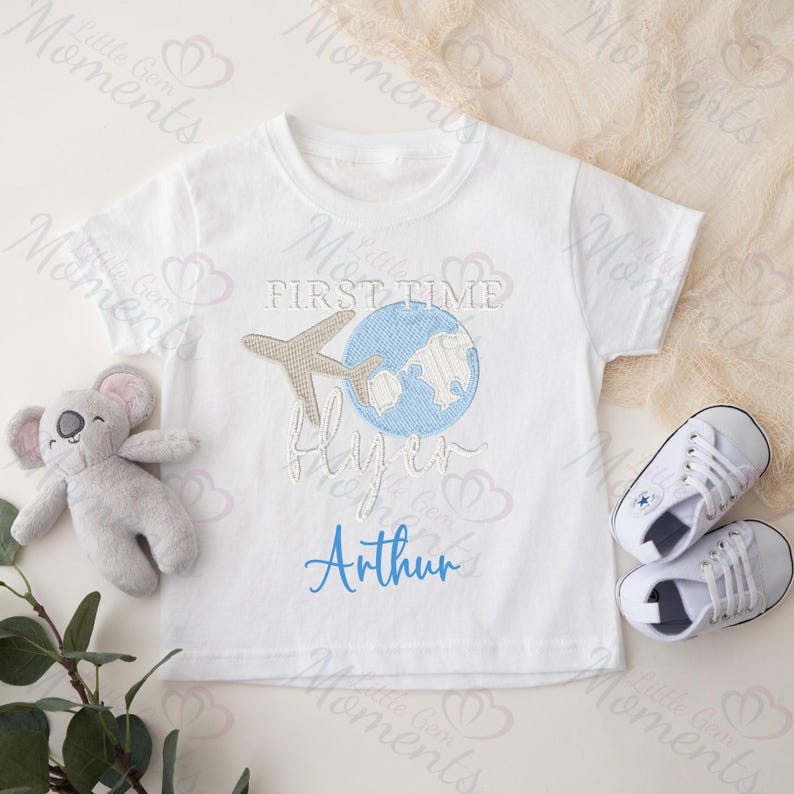 May include: White baby onesie with blue and brown embroidery. The embroidery depicts a globe, airplane, and the text "My First Time Flying In Arthur".