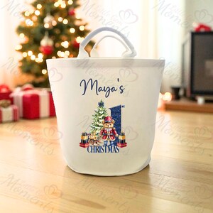Puede incluir: Una bolsa de lona blanca con asa, con el nombre "Maya's" en escritura azul marino. El diseño incluye un árbol de Navidad, un oso de peluche y el texto "CHRISTMAS". La bolsa mide aproximadamente 25 cm de alto.