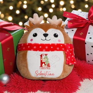 May include: A soft reindeer toy with a red scarf and a white belly. The belly has a Christmas design with a reindeer, tree, and the name "Sebastian". Gifts wrapped in green and white surround the toy.