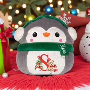 May include: A grey plush penguin toy with a green scarf and hat, and ear muffs. The penguin has a white belly with a Christmas tree, snowman, teddy bear, and the letter "S". The scarf says "Stevie".