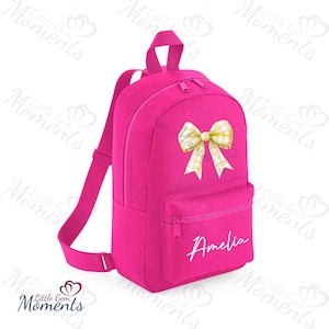May include: A vibrant pink backpack featuring a yellow and white checked bow. The bag has a front zipped pocket and the name "Amelia" in white script. It includes a top handle and adjustable shoulder straps. The bag is suitable for children.
