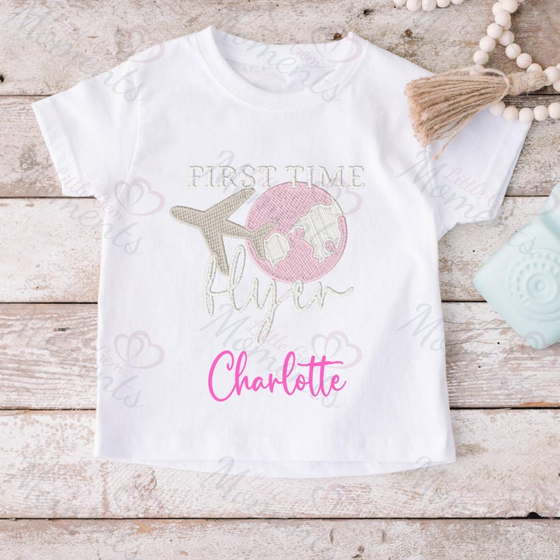 May include: White t-shirt with pink and brown embroidery of a plane flying around a globe. The text "First Time Flyer" is embroidered in pink and the name "Charlotte" is embroidered in pink below.