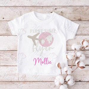 May include: White t-shirt with pink and brown embroidery of a plane and globe. The text "First Time Flyer" is embroidered in pink. The name "Mollie" is embroidered in pink below the design.