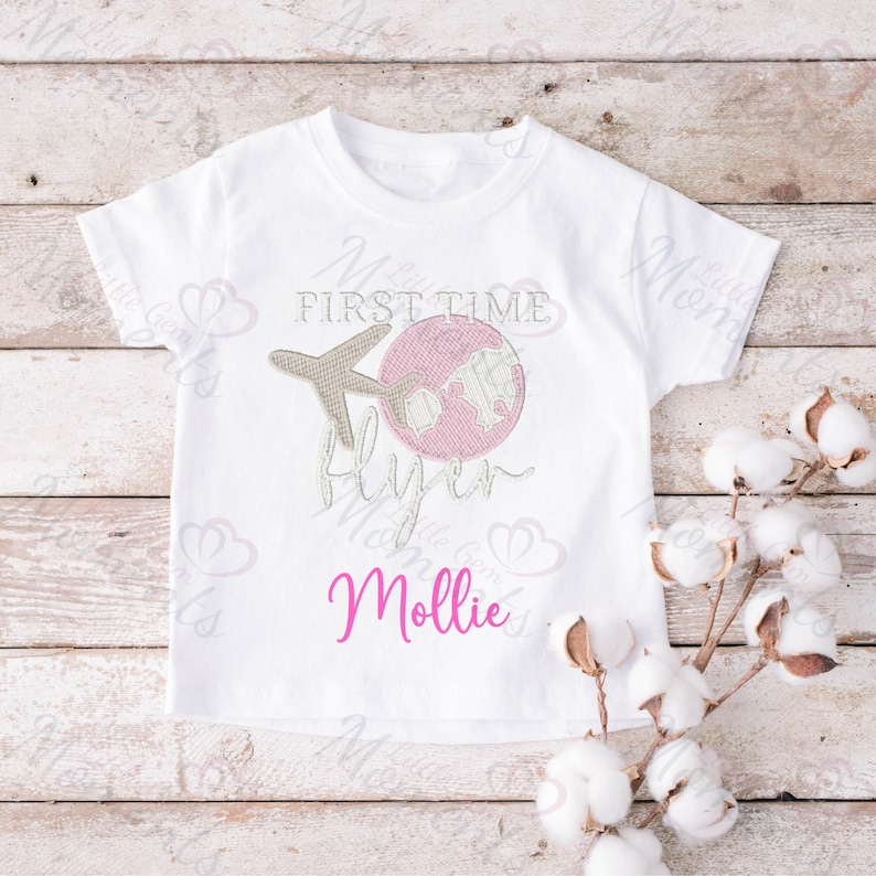 May include: White t-shirt with pink and brown embroidery of a plane and globe. The text "First Time Flyer" is embroidered in pink. The name "Mollie" is embroidered in pink below the design.