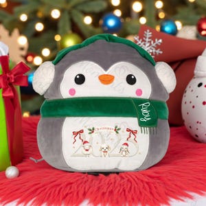 May include: A plush grey penguin with a green scarf and hat, and white earmuffs. The penguin has the name "Ruby" on the scarf and an embroidered design with the text "Christmas 2023" and three cartoon animals. The penguin is on a red surface.