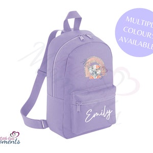 Könnte beinhalten: Lilafarbener Rucksack mit einem Regenbogen und zwei Elefanten, die auf die Vorderseite gestickt sind. Der Rucksack hat eine Reißverschlusstasche auf der Vorderseite und der Name "Emily" ist unter der Tasche gestickt.