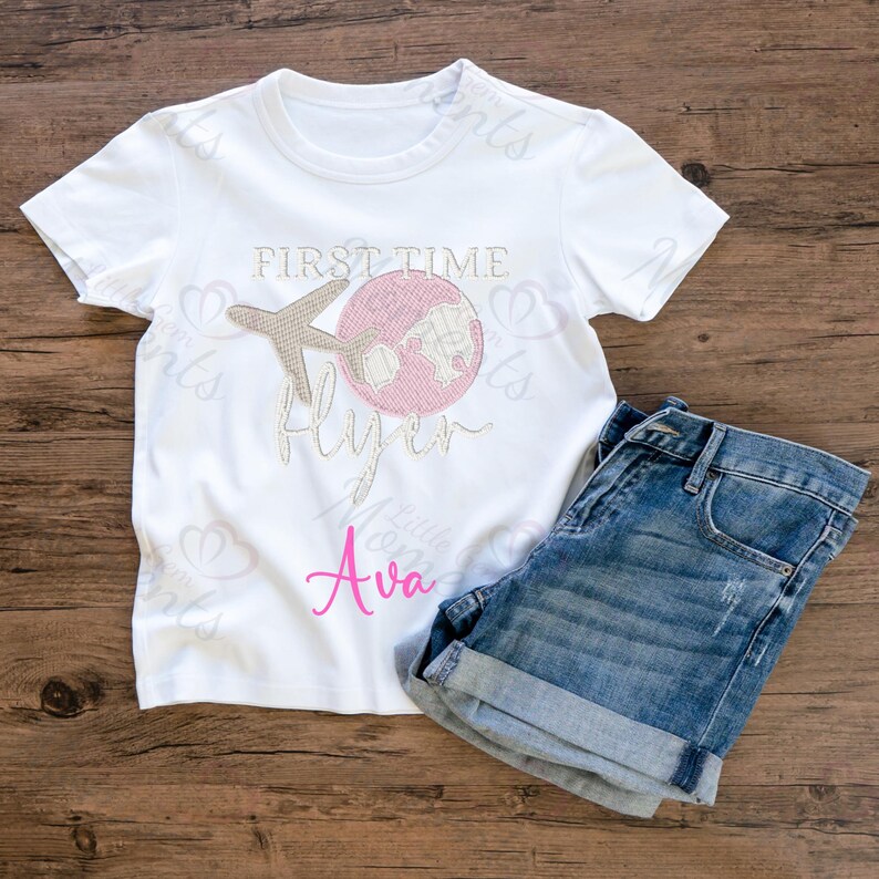 May include: White t-shirt with a pink and brown graphic of a plane flying over a globe. The text "First Time Flyer" is written in brown and the name "Ava" is written in pink.