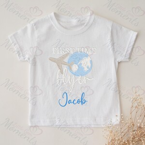 May include: White baby t-shirt with a blue and brown embroidered design of a globe with an airplane flying over it. The text "First Time Flyer" is embroidered in brown and the name "Jacob" is embroidered in blue.