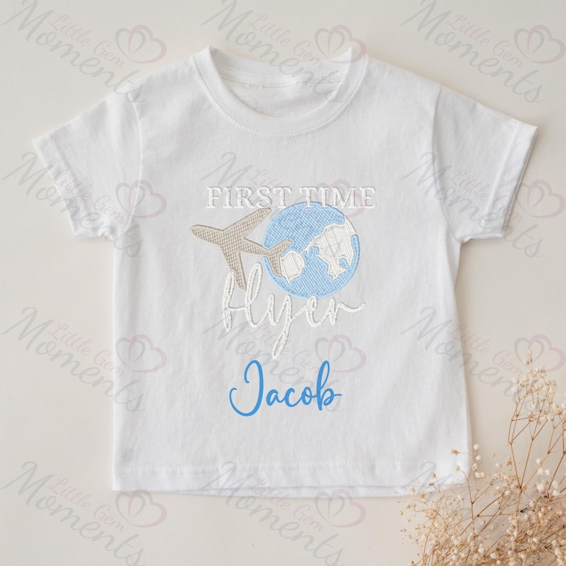 May include: White baby t-shirt with a blue and brown embroidered design of a globe with an airplane flying over it. The text "First Time Flyer" is embroidered in brown and the name "Jacob" is embroidered in blue.