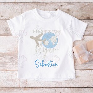 May include: White t-shirt with a blue globe and airplane graphic. The text "My First Time Flying" and the name "Sebastian" are embroidered on the shirt.