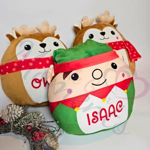 May include: Three plush Christmas-themed cushions. One is a brown reindeer with a red and white scarf and the name "Owen" embroidered on it. Another is a green elf with a red hat and a white shirt with the name "Isaac" embroidered on it. The third is a brown reindeer with a red and white scarf.