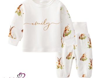 May include: White baby pajama set with a long-sleeved top and matching pants. The set features a repeating pattern of brown bunnies and yellow flowers. The top is personalized with the name "emily" in gold script.