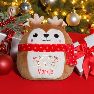 May include: A soft reindeer toy with antlers and a red scarf, featuring a Christmas 2023 design and the name "Marnie". The plush toy is set against a festive backdrop of Christmas decorations and wrapped presents.