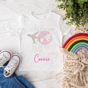 May include: White t-shirt with a pink and gray airplane and globe design. The text "First Time Flyer" is above the design and the name "Connie" is below.