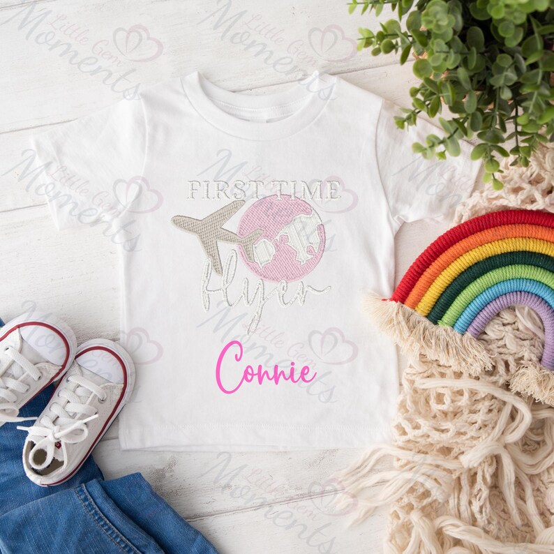 May include: White t-shirt with a pink and gray airplane and globe design. The text "First Time Flyer" is above the design and the name "Connie" is below.