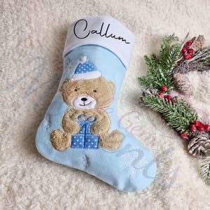 Può includere: Un calzino di Natale blu con un polsino bianco su cui è scritto "Callum". Il calzino presenta un orsacchiotto ricamato che tiene una scatola regalo blu e bianca a pois. Il calzino è decorato con fiocchi di neve bianchi.