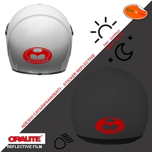 May include: Two motorcycle helmets, one white and one black, with a red reflective sticker of a stylized figure with the text "BUTTMAN". The sticker is reflective and changes color in the light. The helmets are labeled "ADESIVI CATARIFRANGENTI - STICKER REFLECTIVE" and "ORALITE REFLECTIVE FILM".