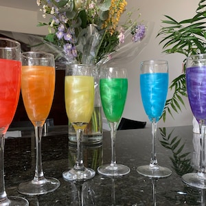 May include: Six champagne flutes filled with colourful iridescent liquid. The liquids are red, orange, yellow, green, blue, and purple. The flutes are arranged in a row on a black surface.