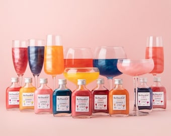 Op de afbeelding: Een verzameling van tien kleine glazen flesjes met kleurrijke vloeistoffen en de tekst 'BeDazzled' en 'Shimmer Summer Syrup' op de etiketten. De flesjes zijn in een rij op een roze oppervlak gerangschikt. Er zijn ook verschillende glazen gevuld met kleurrijke vloeistoffen op de achtergrond.