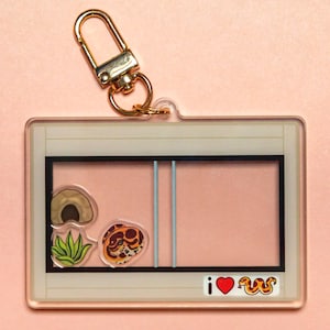 Puede incluir: Un llavero de acrílico transparente con un cierre de metal dorado. El llavero presenta una imagen de dibujos animados de una serpiente dentro de un terrario con una escondite, una planta y el texto "i ♥ s".