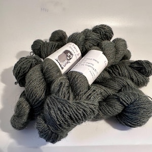 May include: A pile of dark gray yarn skeins with white labels. The labels feature a cartoon drawing of a creature and text that reads "Oviut Blend" and "3 ply fingering". The yarn is a soft, textured material.