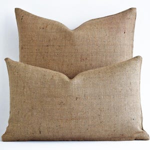 May include: Two rectangular burlap pillows in varying sizes are stacked against a white background. The pillows are a natural tan color, with a coarse, woven texture. The larger pillow is behind the smaller one, creating a layered effect. The pillows are designed for home decor.