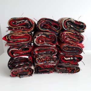 Può includere: Una pila di ritagli di tessuto arrotolati in vari colori, tra cui rosso, nero e marrone. Il tessuto presenta un mix di trame e motivi, con bordi visibili e un aspetto stratificato. I rotoli sono ben disposti.