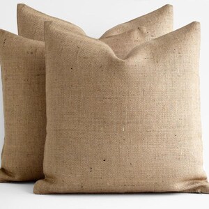 May include: Two square throw pillows made of a coarse, tan-colored burlap fabric. The pillows feature a visible woven texture with natural variations in the fiber color, creating a neutral appearance. Each pillow has a simple, clean-edged design.