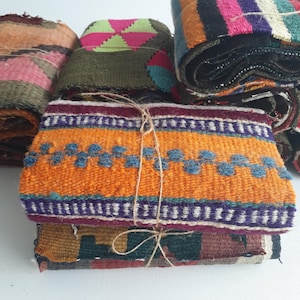 May include: A stack of colorful woven fabric pieces, tied with twine. The fabrics display various patterns and colors, including orange, blue, purple, pink, and green. The textiles appear to be hand-woven, with visible textures and details.