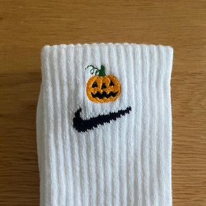 May include: A white ribbed crew sock featuring an embroidered orange jack-o'-lantern pumpkin with a green stem positioned directly above a black Nike swoosh logo. The sock is made of a textured knit fabric and displays a clean, folded design.
