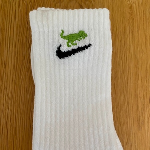 May include: White cotton socks with a black Nike swoosh logo and a green dinosaur graphic.