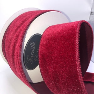 Könnte beinhalten: Eine Spule burgunderfarbenes Samtband mit glitzerndem Finish. Das Band ist 2,5 Zoll breit und hat einen drahtverstärkten Rand.
