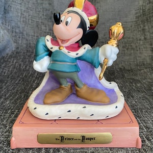 May include: A porcelain figurine of Mickey Mouse dressed as a prince from the Disney movie "The Prince and the Pauper". He is wearing a purple and gold robe, a crown, and holding a scepter. The figurine is on a pink base with a gold plaque that reads "The Prince and the Pauper".