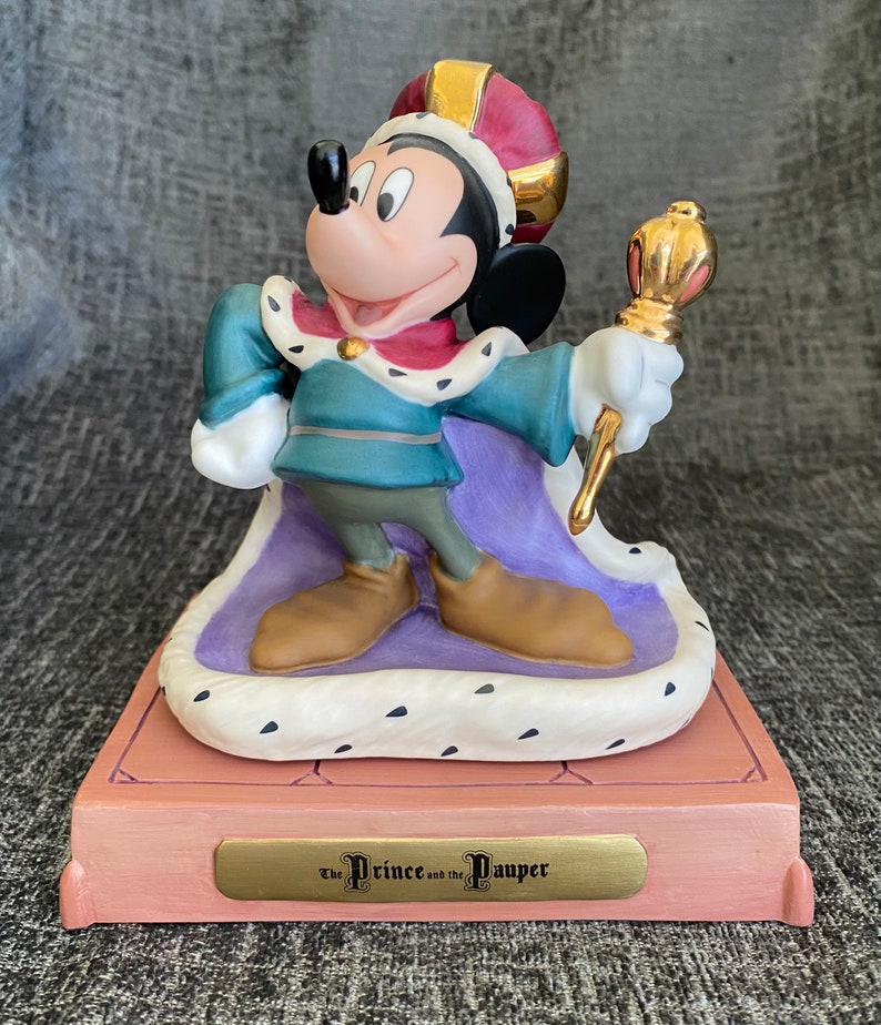 May include: A porcelain figurine of Mickey Mouse dressed as a prince from the Disney movie "The Prince and the Pauper". He is wearing a purple and gold robe, a crown, and holding a scepter. The figurine is on a pink base with a gold plaque that reads "The Prince and the Pauper".