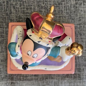 May include: A ceramic figurine of Mickey Mouse dressed as a king. He is wearing a gold crown with red jewels, a white and gold robe, and a blue and green tunic. He is holding a gold scepter in his right hand. The figurine is sitting on a pink and purple base.