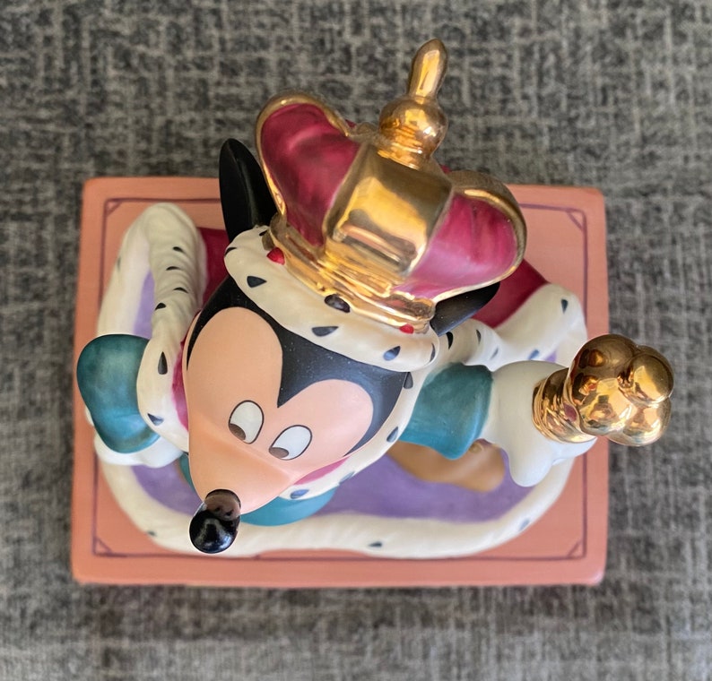 May include: A ceramic figurine of Mickey Mouse dressed as a king. He is wearing a gold crown with red jewels, a white and gold robe, and a blue and green tunic. He is holding a gold scepter in his right hand. The figurine is sitting on a pink and purple base.
