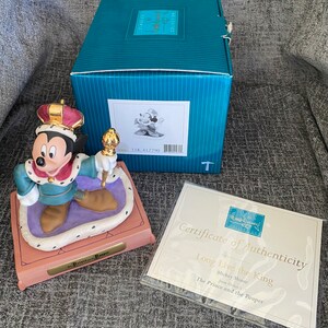 May include: A ceramic figurine of Mickey Mouse dressed as a king from Disney's *The Prince and the Pauper*. Mickey is wearing a gold crown, a blue and gold robe, and holding a golden scepter. The figurine is on a purple and gold base with the words "The Prince and the Pauper" inscribed on it. The figurine comes with a certificate of authenticity.