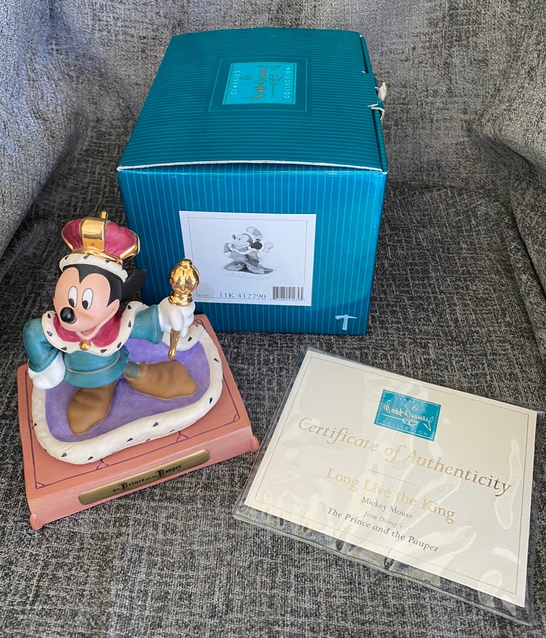 May include: A ceramic figurine of Mickey Mouse dressed as a king from Disney's *The Prince and the Pauper*. Mickey is wearing a gold crown, a blue and gold robe, and holding a golden scepter. The figurine is on a purple and gold base with the words "The Prince and the Pauper" inscribed on it. The figurine comes with a certificate of authenticity.