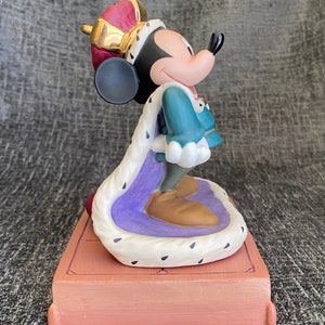 May include: A porcelain figurine of Mickey Mouse dressed as a king. He is wearing a purple and white robe with a gold crown and a blue and green tunic. He is standing on a brown platform.