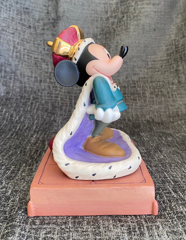 May include: A porcelain figurine of Mickey Mouse dressed as a king. He is wearing a purple and white robe with a gold crown and a blue and green tunic. He is standing on a brown platform.