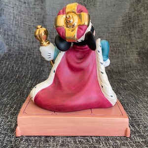 May include: A porcelain figurine of Mickey Mouse dressed as a king. He is wearing a red and white robe, a gold crown, and holding a scepter. He is standing on a platform with a gold trim.