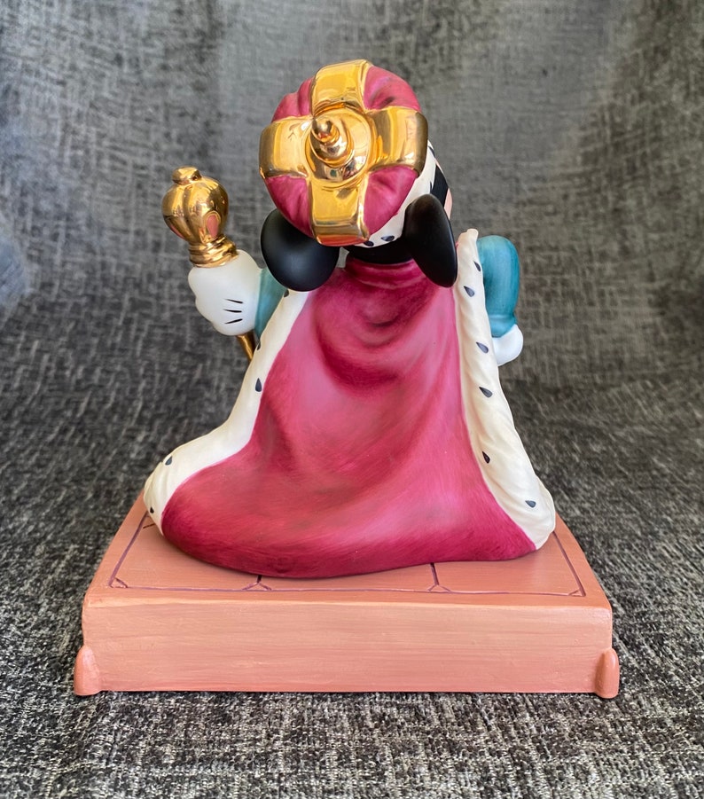 May include: A porcelain figurine of Mickey Mouse dressed as a king. He is wearing a red and white robe, a gold crown, and holding a scepter. He is standing on a platform with a gold trim.