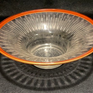 May include: A clear glass bowl with a wide, shallow base and a decorative, etched pattern. The bowl has a red rim with a thin gold band.