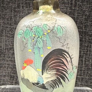 May include: A small, clear glass bottle with a silver and turquoise stopper. The bottle is decorated with a hand-painted image of a rooster and a green plant.