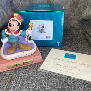 May include: A ceramic figurine of Mickey Mouse as a king from Disney's The Prince and the Pauper. Mickey is wearing a gold crown, a blue and green robe, and gold shoes. He is holding a gold scepter. The figurine is on a pink and purple base with a gold plaque that reads "The Prince and the Pauper".