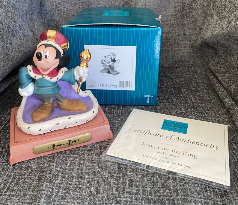 May include: A ceramic figurine of Mickey Mouse as a king from Disney's The Prince and the Pauper. Mickey is wearing a gold crown, a blue and green robe, and gold shoes. He is holding a gold scepter. The figurine is on a pink and purple base with a gold plaque that reads "The Prince and the Pauper".